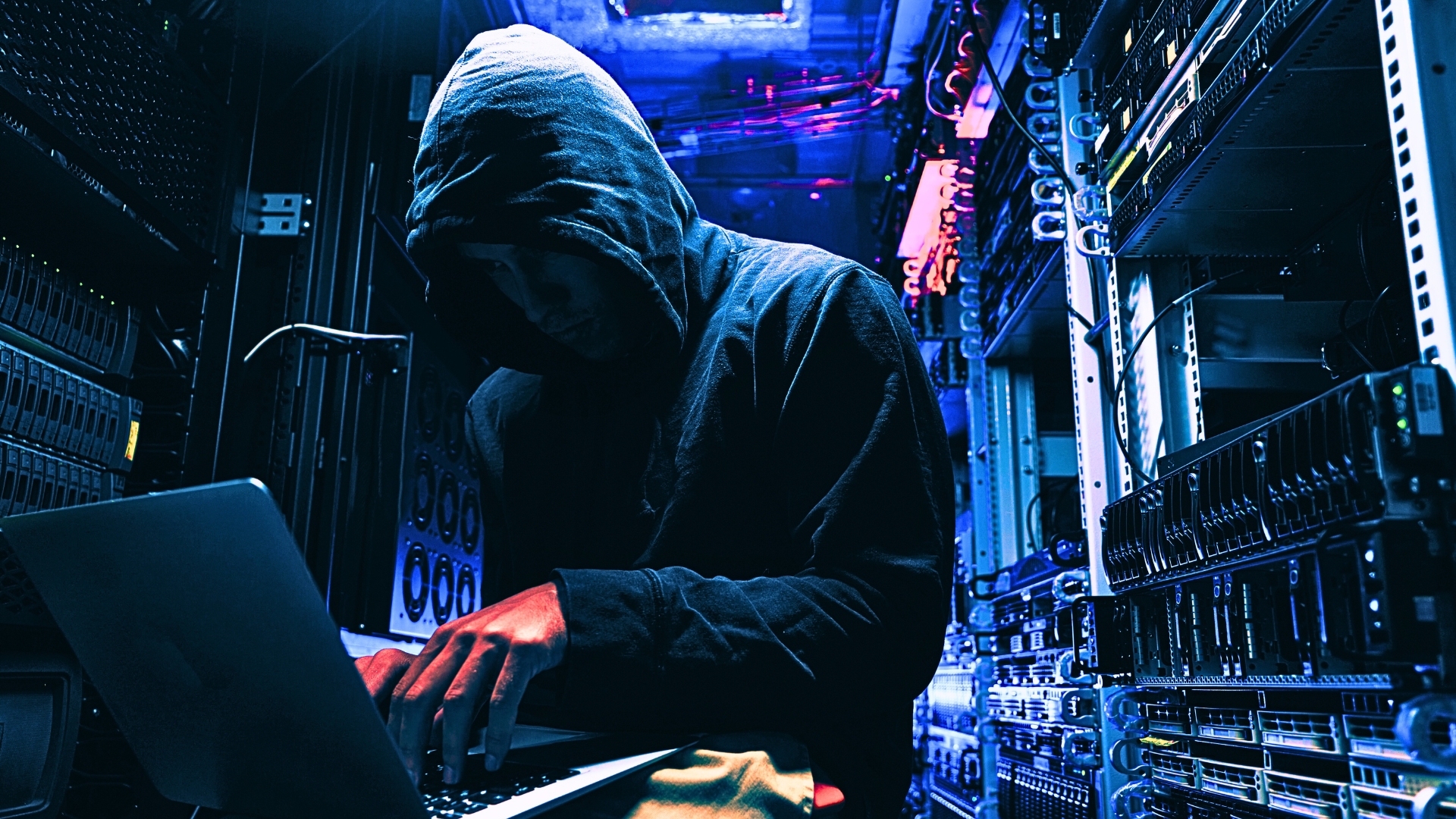 suspicious hooded figure sitting at a laptop in a server room cybersecurity cyberattack awareness