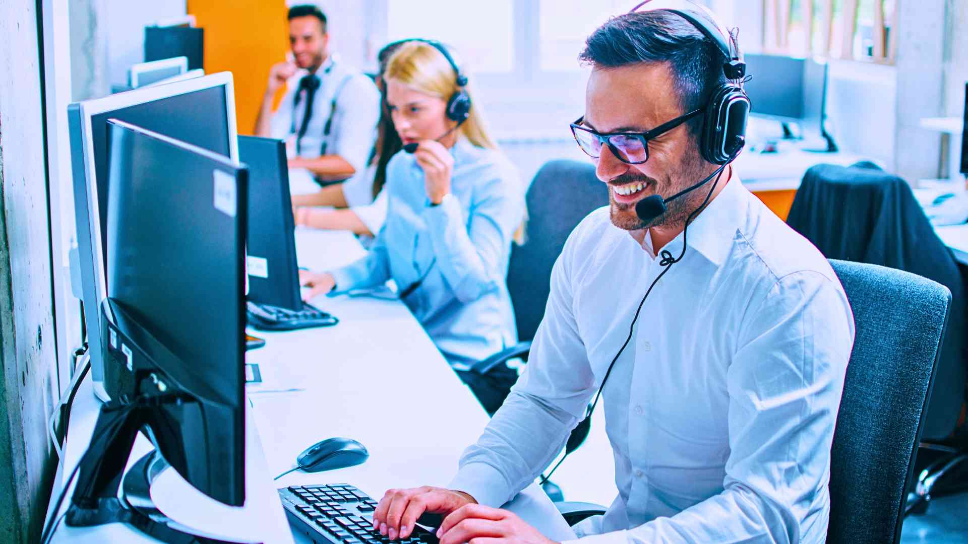managed service provider worker sitting at desk with coworkers nearby typing on keyboard talking on headset on computer
