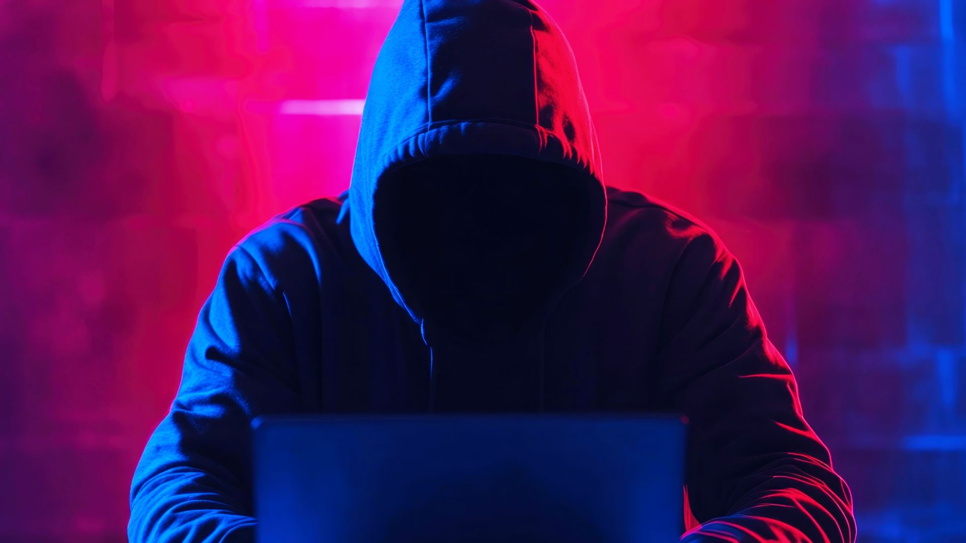hooded figure leaning over and typing on a laptop with face hidden and red danger background