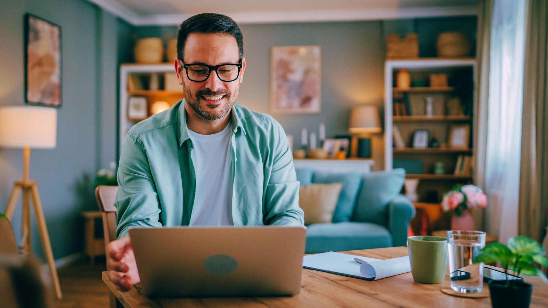 male wearing glasses typing on laptop at remote work home office setup with coffee and glass of water next to them