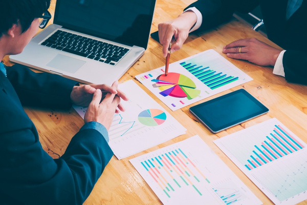 business people pointing at sheet of paper with pie chart and graphs on it next to a laptop and tablet