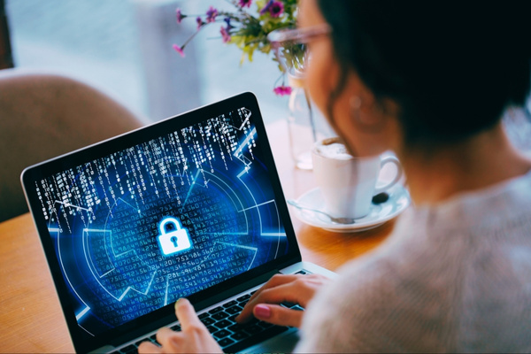 female business person sitting at desk typing on their laptop keyboard with a blue cybersecurity screen showing
