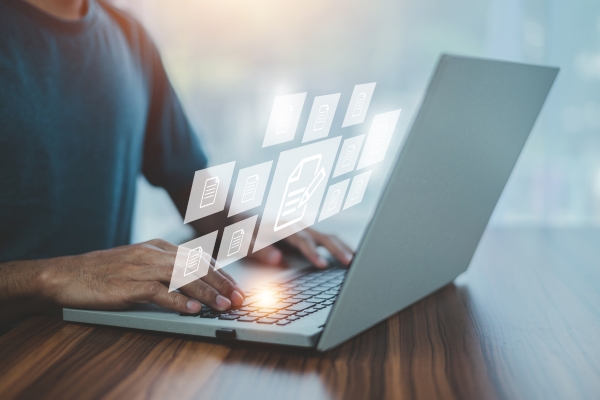 person typing on a laptop keyboard on a wooden desk with document icons hovering above