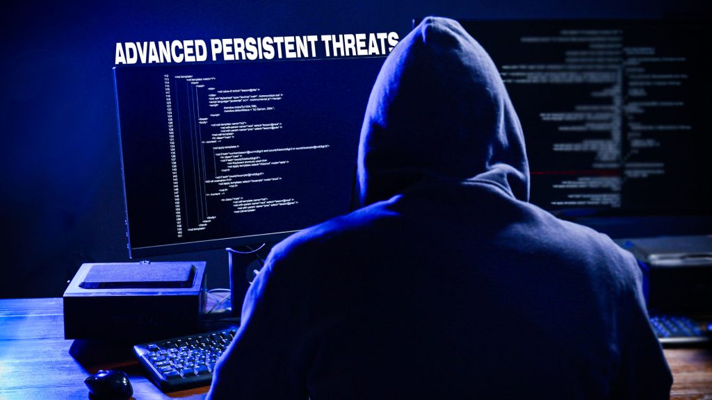 cybercriminal sitting at desk wearing a hoodie typing on keyboard performing an advanced persistent threat cyberattack